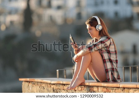 Side view portrait of a happy teenage girl listening to music sitting on a ledge in a town outskirts
