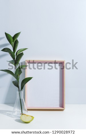 Mock up white frame, green twigs in vase, piece of apple and glass of limonade on book shelf or desk. White colors. White-blue colors. Minimalistic concept.