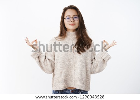 Girl keeps patience, being calm and peaceful as posing in sweater and glasses looking at camera, smiling standing in lotus pose with mudra gesture, meditating or doing yoga for relaxation of mind