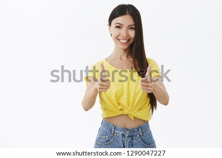 Friendly attractive and outgoing supportive tanned female with acne and natural beauty without makeup showing thumbs up and smiling broadly approving and liking awesome idea against grey wall