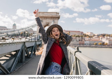 Blissful pale woman in coat expressing true emotions while posing on bridge in warm day. Pleased white lady with light-brown curls dancing near river on city background.