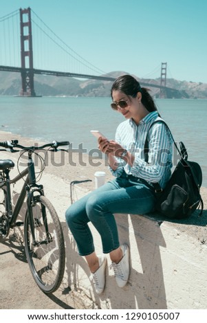 asian female college student looking in mobile phone touching screen and pausing on bay along the coast by riding bike. young happy girl texting message on cellphone sitting relaxing under sunlight.