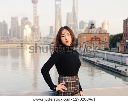 Beautiful young brunette woman in black clothes posing with blur Shanghai Bund landmark buildings background in autumn dusk light.  Emotions, people, beauty, travel and lifestyle concept.
