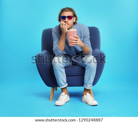 Emotional man with 3D glasses and popcorn sitting in armchair during cinema show on color background