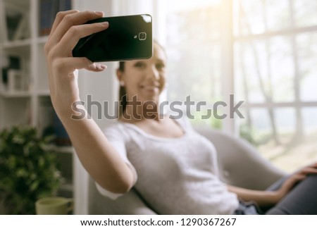 Happy girl taking selfies using her smartphone and sitting on the armchair in the living room
