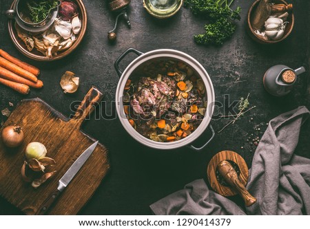 Cooking preparation of stewed meat.  Roasted beef meat in cast iron cooking pot with vegetables on dark rustic background with kitchen utensils and seasoning, top view with copy space. 