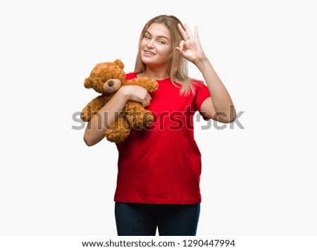 Young caucasian woman holding cute teddy bear over isolated background doing ok sign with fingers, excellent symbol