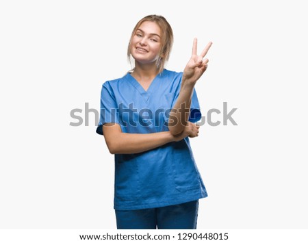 Young caucasian nurse woman wearing surgeon uniform over isolated background smiling with happy face winking at the camera doing victory sign. Number two.