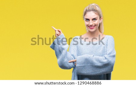 Young beautiful blonde and blue eyes woman wearing winter sweater over isolated background with a big smile on face, pointing with hand and finger to the side looking at the camera.