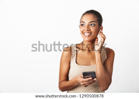Beautiful young african woman casually dressed standing isolated over white background, wearing wireless earphones, using mobile phone