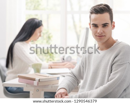 Handsome confident student posing and smiling, a girl is using a computer in the background, learning and education concept