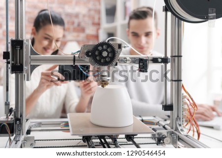 Engineering students printing prototype models using a 3D printer, the girl is taking pictures with a smartphone and sharing online