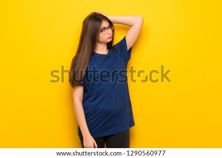 Young woman with glasses over yellow wall having doubts while scratching head