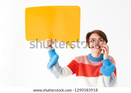 Happy cheerful girl wearing sweater standing isolated over white background, holding empty speech bubble, talking on mobile phone