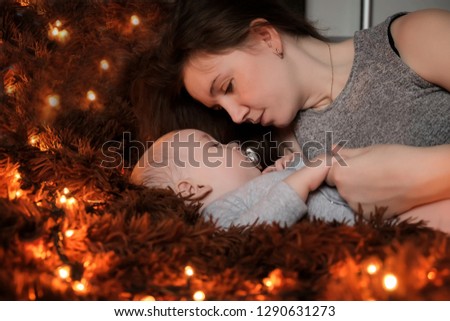 A young mother looks at her baby who sleeps on a fluffy brown blanket, lights are spread around them.