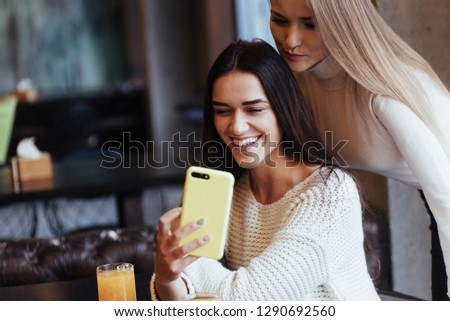 Let's check our photo. In the restaurant. Two female friends sit indoors with yellow drink and use the smartphone.