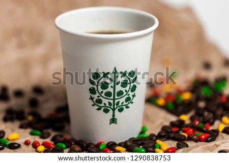 Paper glass with a picture and a drink. Coffee beans, cinnamon and glazed sweets are next to each other on the table. Brown tones in the photo.