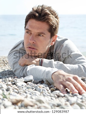 Close up portrait of a man laying down on the shore of a white pebble beach being thoughtful with the blue sea in the background.