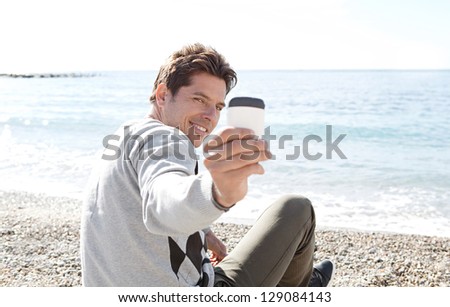 Portrait of a smart man sitting on a pebble beach, turning around to take a picture with his "smart phone", smiling.