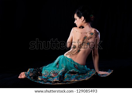 Low key profile portrait of beautiful, brunette woman sitting on the ground with her back to the camera. She has a peacock feather henna design on her back. Shot in the studio on a black background.