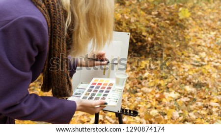 Close-up of young blonde girl artist in purple coat that paints a picture on the easel in the autumn park, holding brush and paints in hands. Autumn concept.