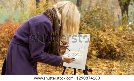 Close-up of young blonde girl artist in purple coat that paints a picture on the easel in the autumn park, holding brush and paints in hands. Autumn concept.