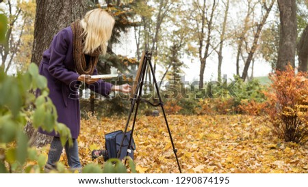 Beautiful blonde girl in the purple coat painting a picture on an easel in the autumn park near big tree, holding paints in hand. Colorful leaves on background