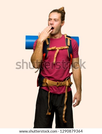 Hiker man with mountain backpacker yawning and covering wide open mouth with hand on isolated background