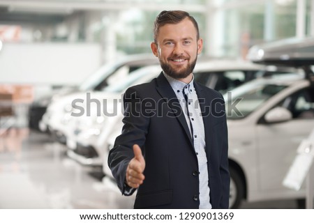 Handsome manager of car dealership looking at camera, smiling, posing. Car dealer wearing in official shirt, dark blue suit. Happy bearded man showing thumb up.