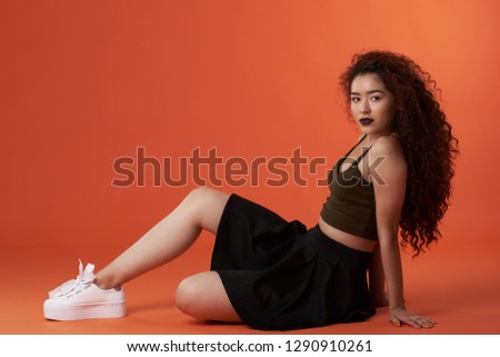 Young hispanic woman with curly hair in sitting pose on orange studio background