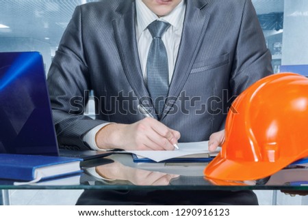 A closeup of an engineer or an owner of a building company signing documents while sitting at the glass table at the office interior background. The concept of successful contract or agreement.