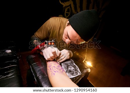 Handsome young guy in a black hat and with tattoos, beats a tattoo on his arm, tattoo salon, tattoo artist Royalty-Free Stock Photo #1290954160