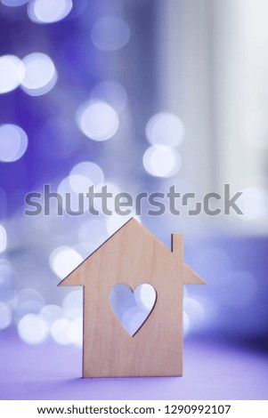 Wooden icon of house with hole in the form of heart on near window in daylight with bokeh lights on blurred purple background. Romantic card with copy space. Concept of sweet home.