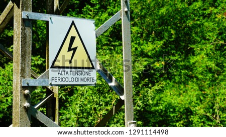 sign with the inscription in Italian HIGH VOLTAGE DANGER OF DEATH on electric pylon among the plants