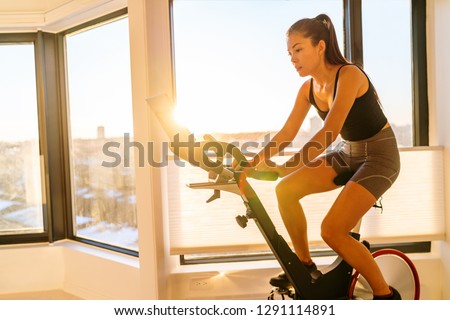 Home fitness workout woman training on smart stationary bike indoors watching screen connected online to live streaming subscription service for biking exercise. Young Asian woman athlete. Royalty-Free Stock Photo #1291114891