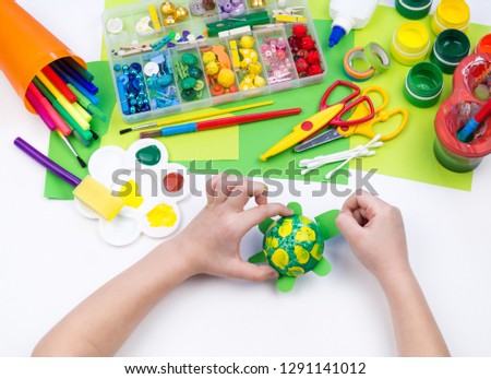 The child makes a craft toy from foam plastic tortoise. Material for creativity and education. Hand kid paints paint. White background.