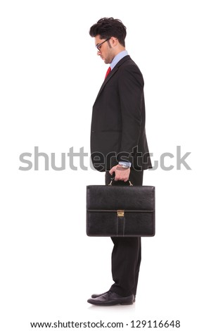 side view full length picture of a young business man standing with his suitcase in his hand and looking down, in front of him on white background