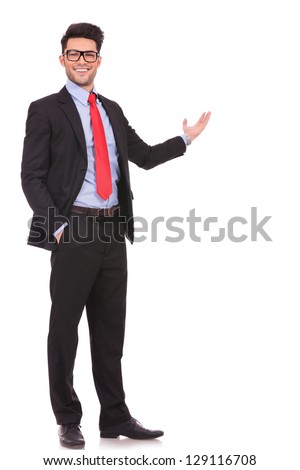full length picture of a young business man presenting something in the back with one hand in his pocket while looking at the camera with a smile on his face, on white background
