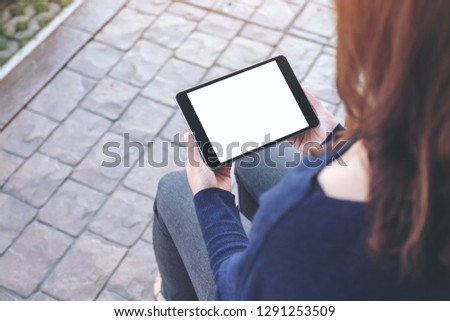 Mockup image of a woman holding black tablet pc with blank white screen horizontally while sitting in the outdoors