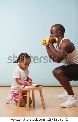Cute little daughter fitness training her handsome black young father. She is making notes in her paper notebook. Child role play. He looks at her for approval, while making squats. Last push.
