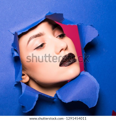 The face of young beautiful girl with a bright make-up and puffy dark lips peers into a hole in blue paper.