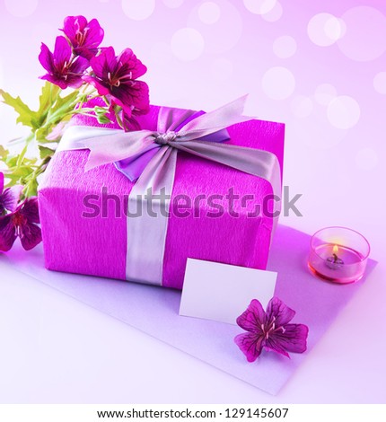 Photo of pink giftbox with silk bow, fresh purple flowers, candle and blank postcard isolated on blur background, festive still life, happy mothers day, romantic holiday, love and romance concept