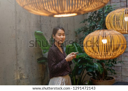 Asian hipster girl is dreaming about something good during her chat in social network via smart phone, young interior blogger take a pictures in cozy coffee shop - Image 