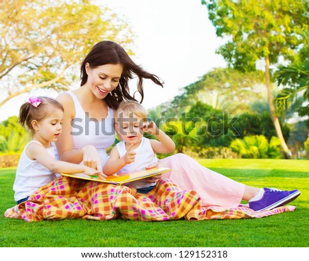 Picture of beautiful woman with daughter and son sitting down on green grass field and read fairytale, cute female with two little children enjoying book outdoors, preschool education, happy family