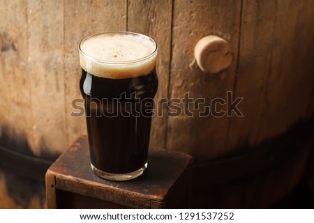 Pint glass of dark stout beer standing near an old wooden barrel in a cellar Royalty-Free Stock Photo #1291537252