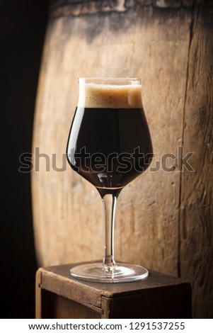 Glass of dark stout beer standing near an old wooden barrel in a cellar Royalty-Free Stock Photo #1291537255