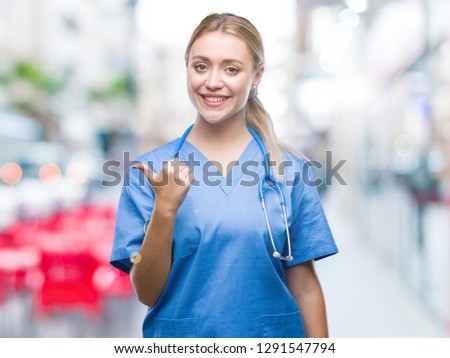 Young blonde surgeon doctor woman over isolated background smiling with happy face looking and pointing to the side with thumb up.