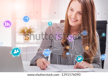 Cheerful young businesswoman writing and looking at her laptop screen in office with colorful business icons in the foreground. Toned image double exposure
