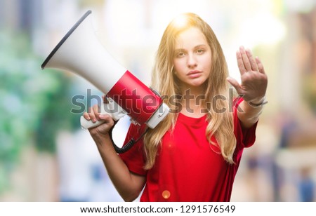 Young beautiful blonde woman yelling through megaphone over isolated background with open hand doing stop sign with serious and confident expression, defense gesture