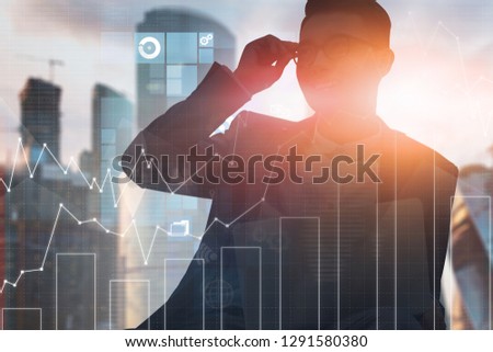 Cheerful Asian businessman in glasses sitting over modern cityscape background with double exposure of graphs and business interface icons. Toned image
